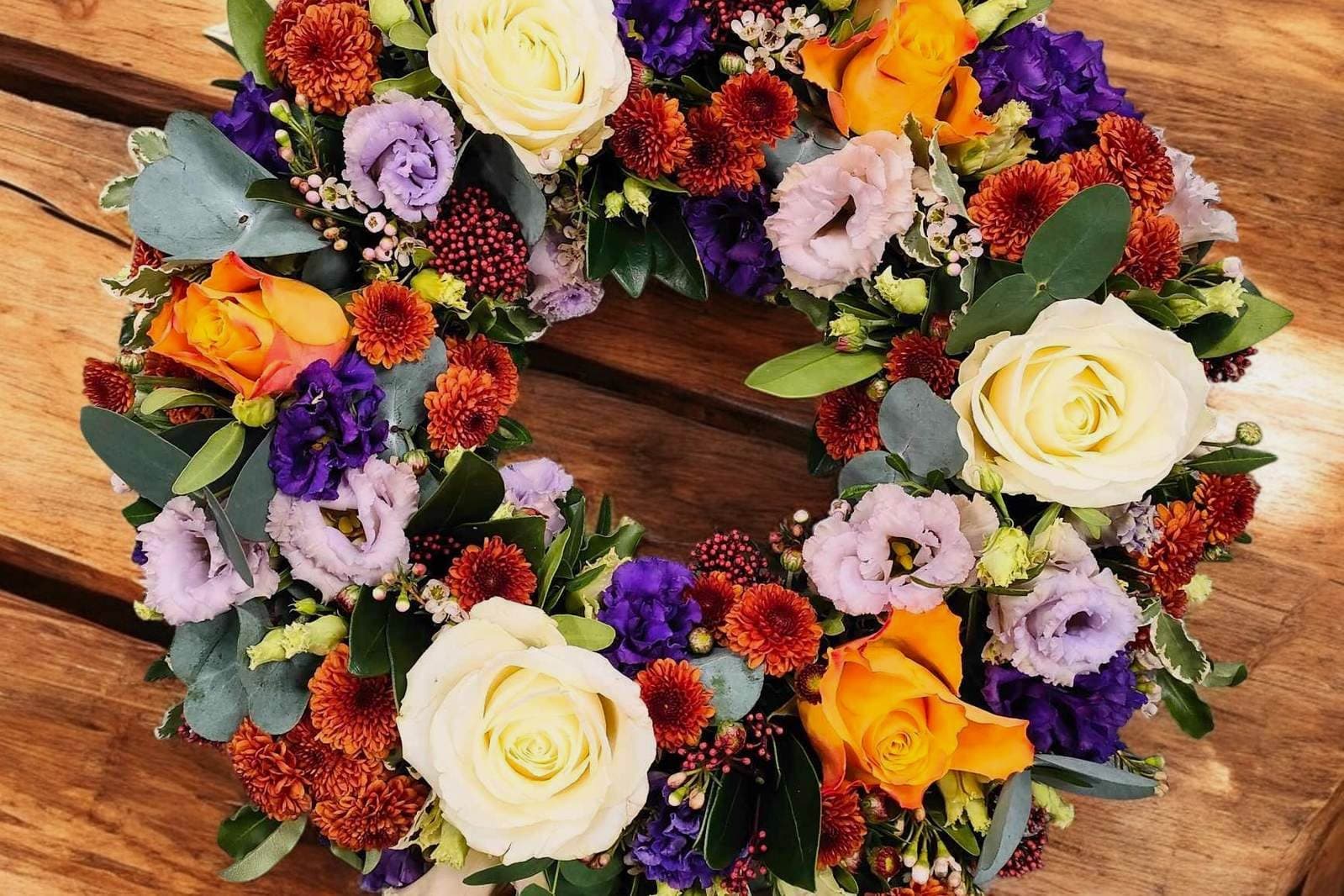 A round funeral tribute wreath of cream and orange roses, purple lisianthus and rust chrysanthemums on a wooden table