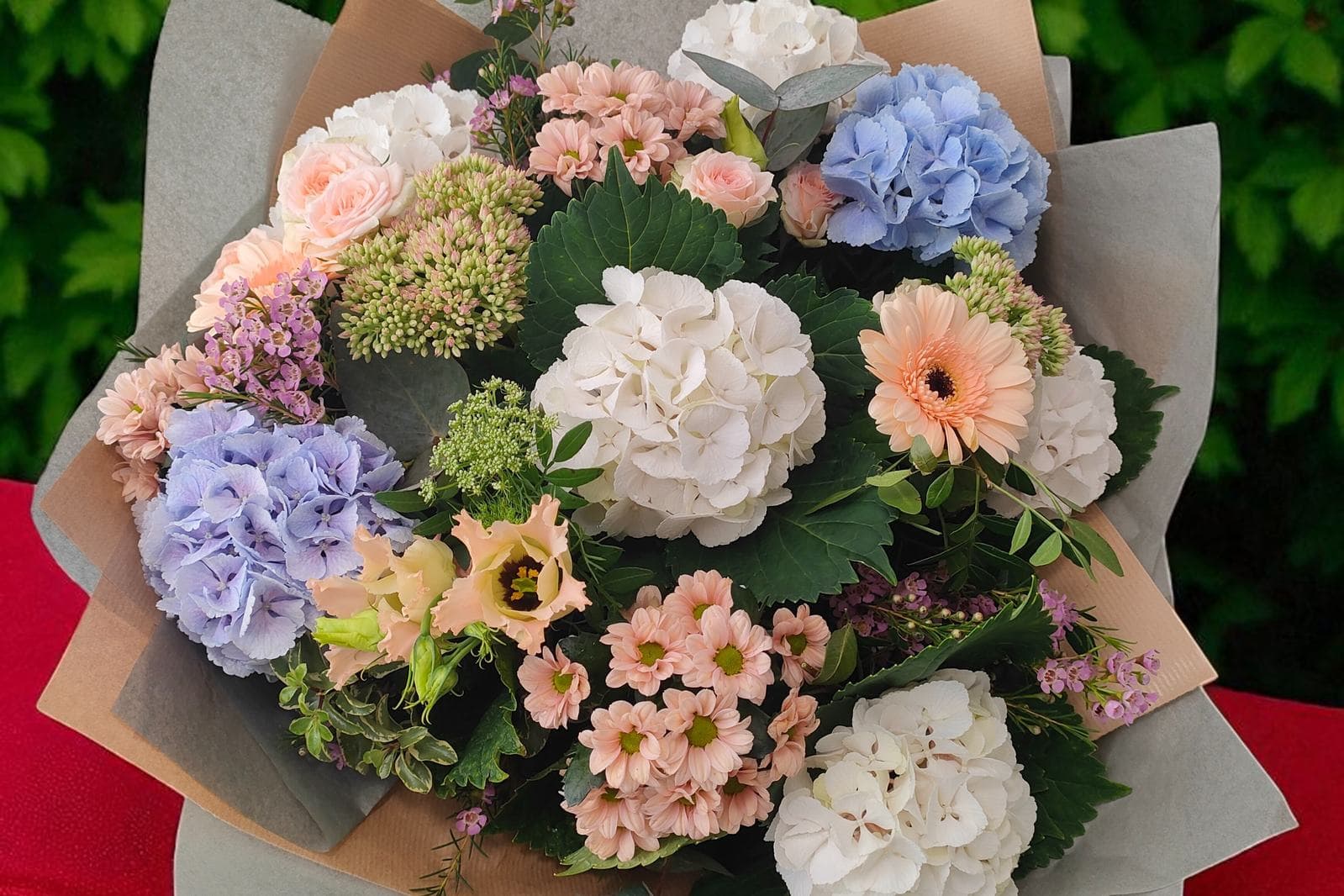 A hand-tied bouquet of white and blue hydrangeas, peach roses, gerbera daisies and pink chrysanthemums wrapped in grey and kraft paper