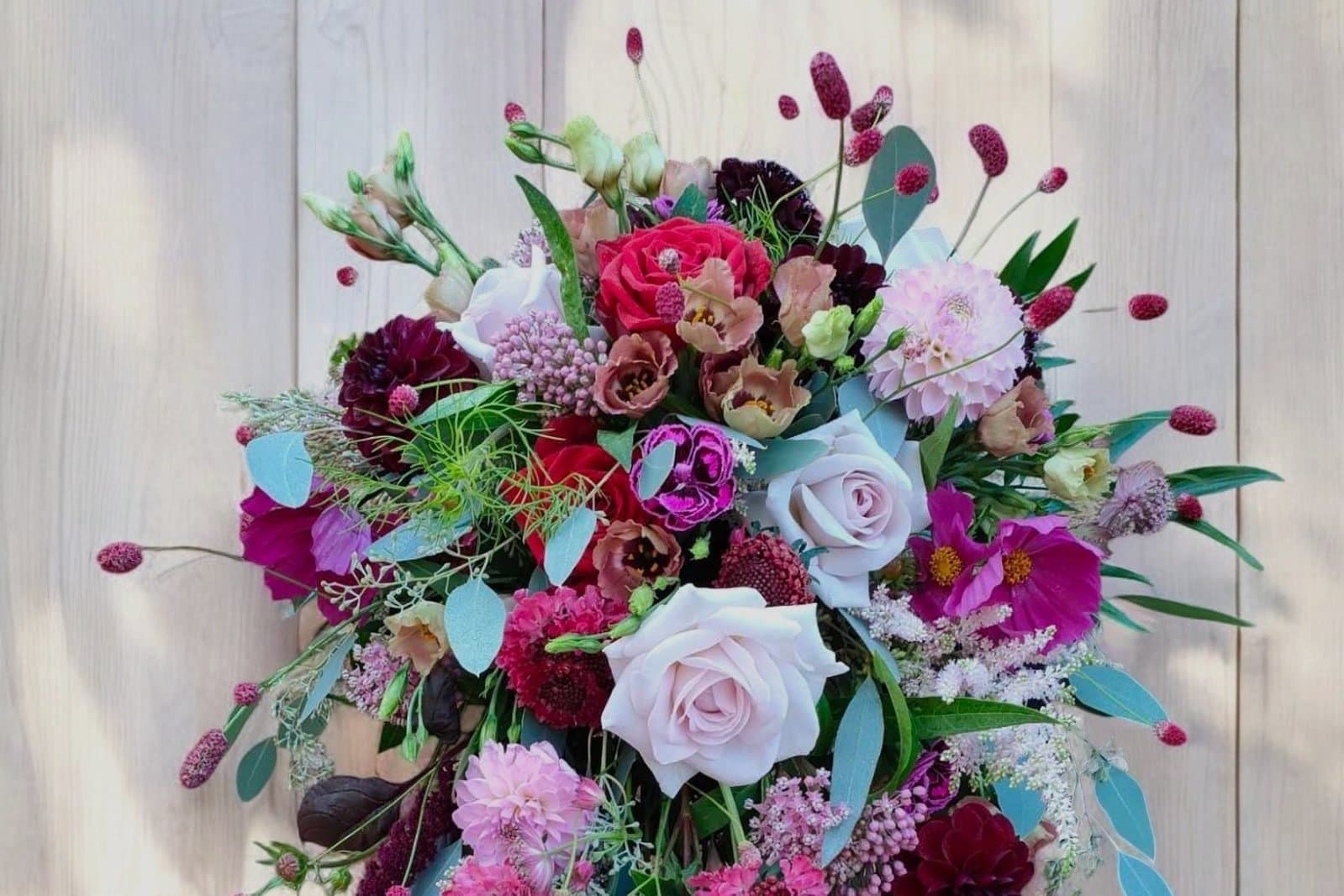 A cascading wedding bouquet of blush and burgundy roses, dahlias, scabious and cosmos against a pale wood-plank backdrop
