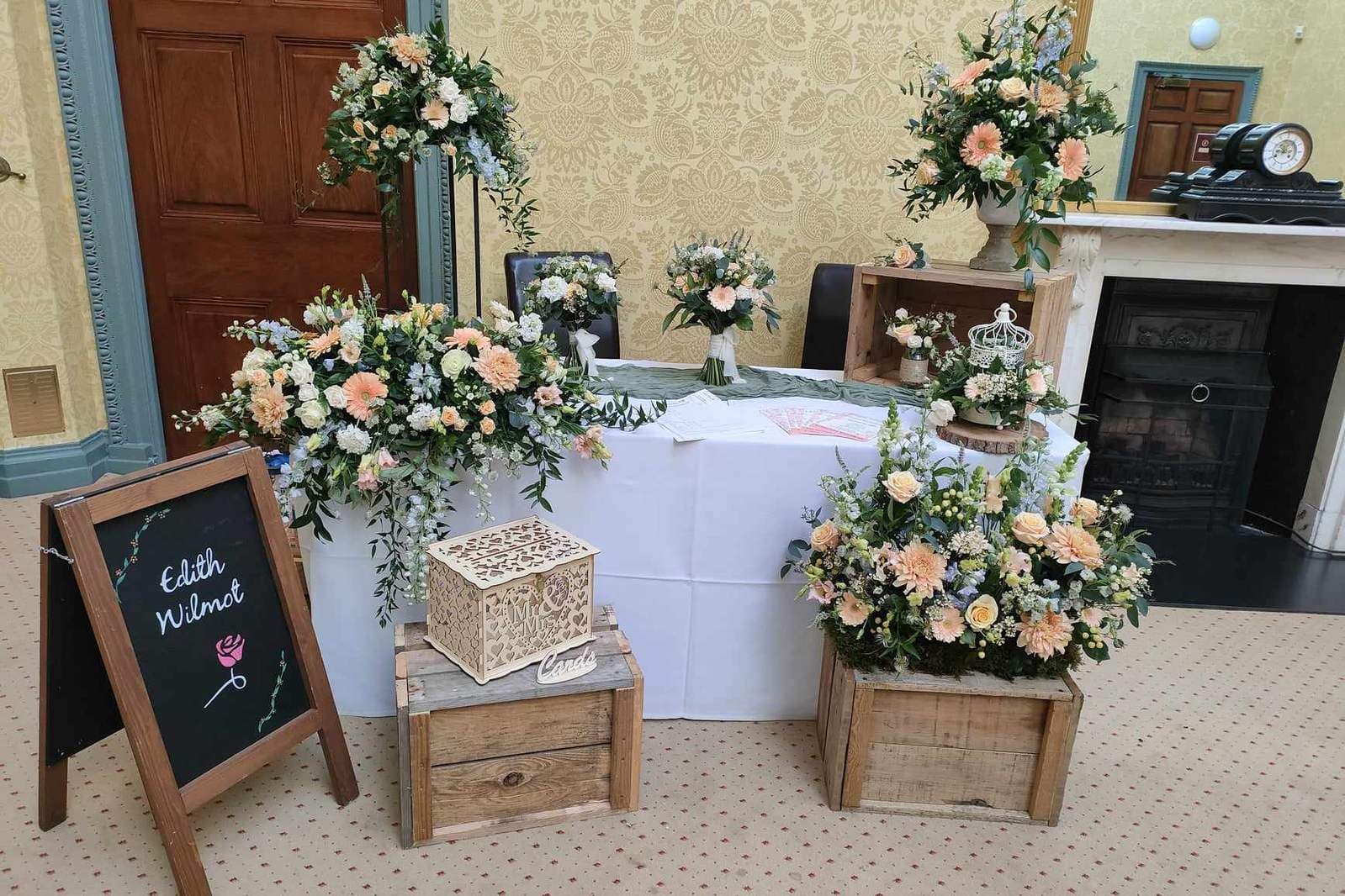 Peach and white wedding florals displayed on crates and a table beside an easel sign reading Edith Wilmot at a reception venue