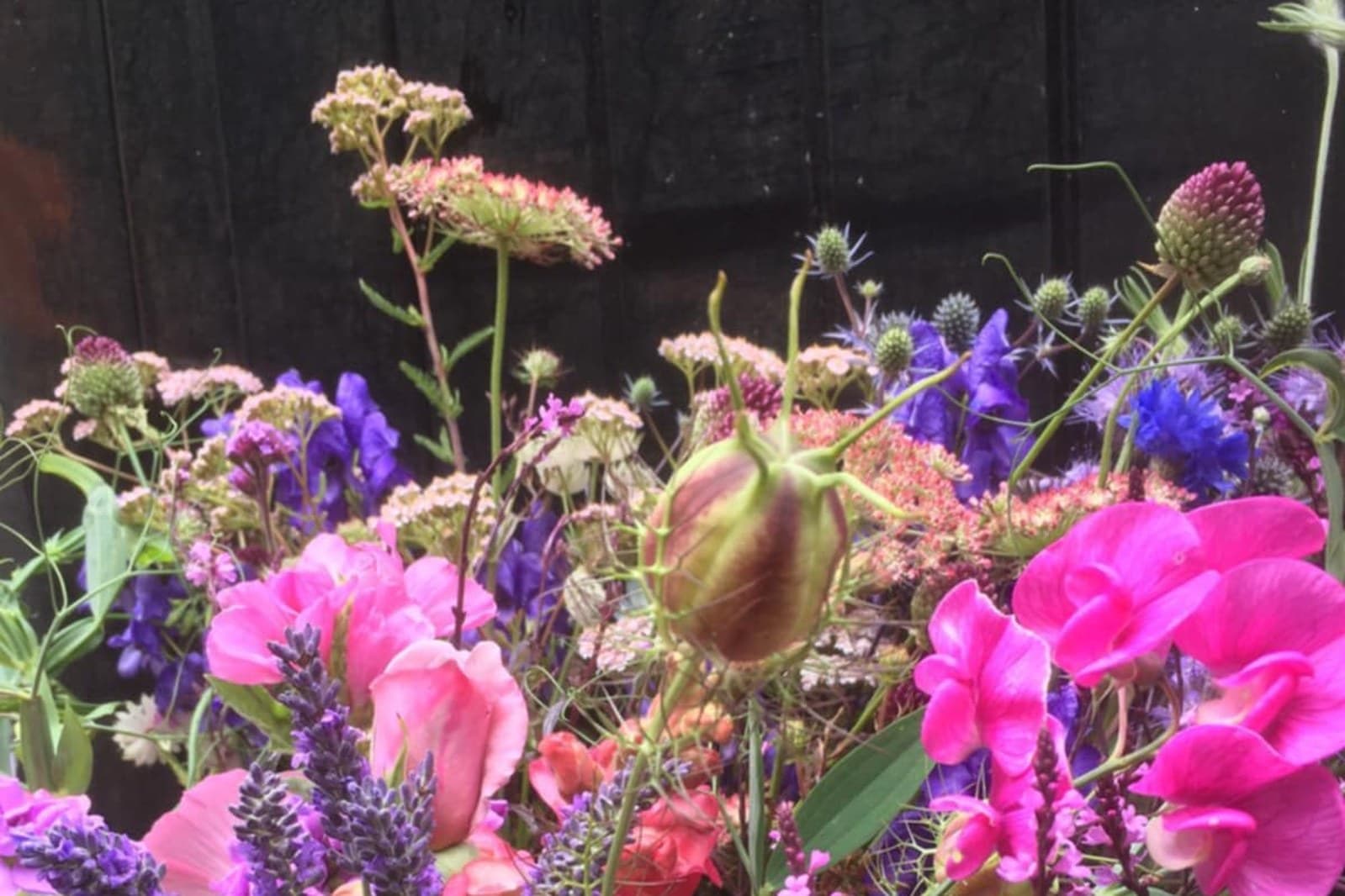 A dense mix of British-grown cut flowers including pink sweet peas, love-in-a-mist, sea holly, cornflowers and yarrow against a dark wooden fence